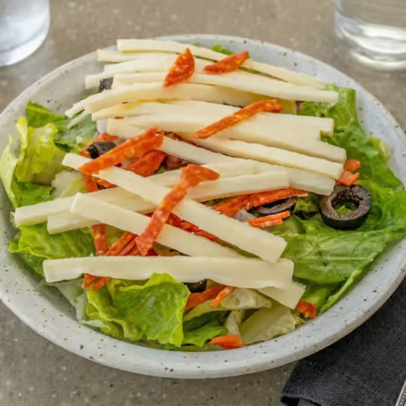 Fresh green salad with white cheese sticks, pepperoni, and black olives in a white bowl.