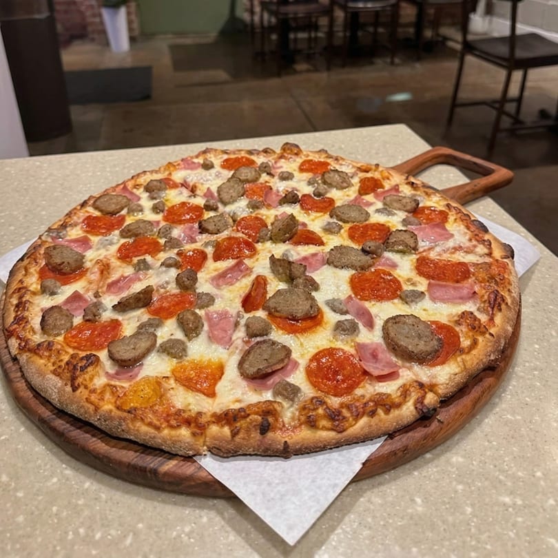 A large, round pizza with pepperoni, sausage, and ham sits on a wooden serving board.